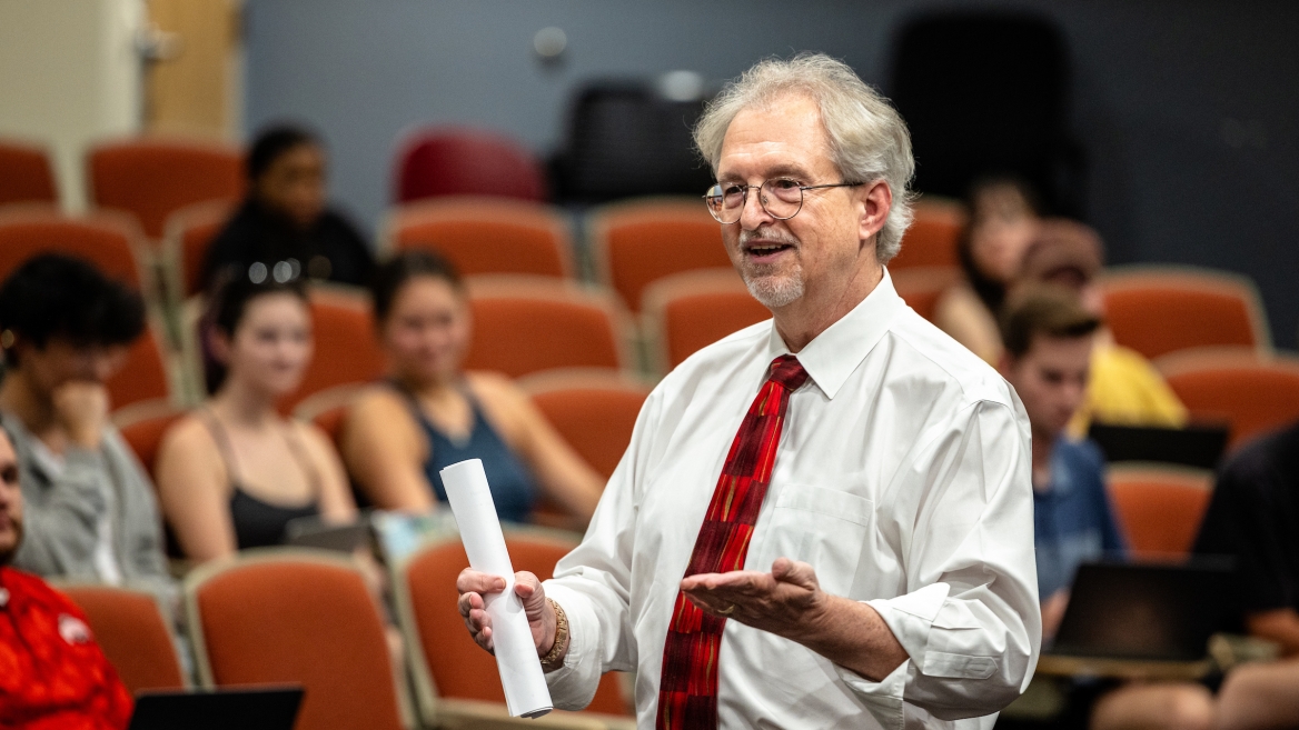 Professor in a white shirt and red tie speaking to students in a classroom.