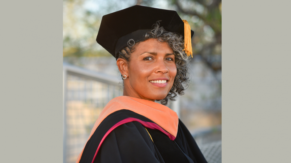 Dawn Augusta smiles at the camera. She is wearing her Doctorate Graduation Regalia complete with black hat, gold tassel and black gown with maroon trimming. 