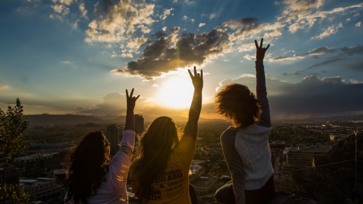 three women pictured from behind as they raise their hands toward a sunset