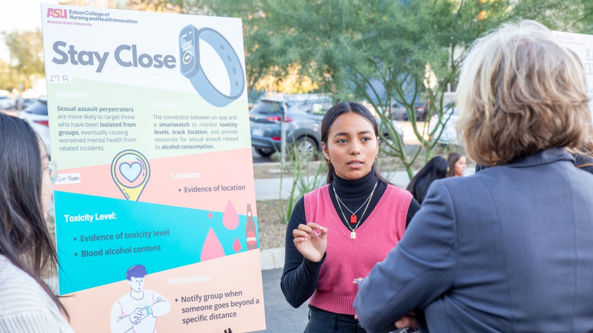 A student presents her team idea to Edson College Dean Judy Karshmer. The student is wearing a bright pink vest over a black turtleneck and is standing in front of a poster with the words "stay close" on it.