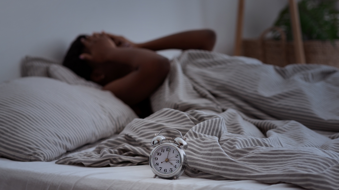 Woman with her hands on her face lying in bed, with an alarm clock next to her.