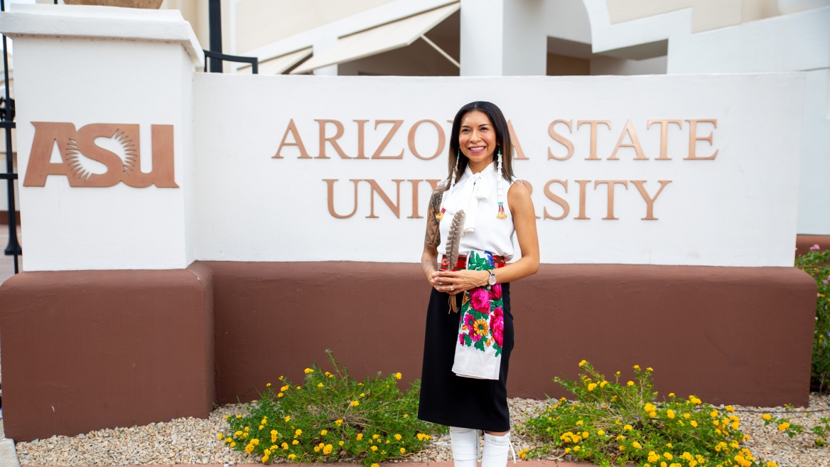 Kristin Payestewa-Picazo stands in front of an Arizona State University sign. She is wearing traditional clothing representing her Hopi and Navajo heritage. She has a feather in between her hands and smiles at the camera.