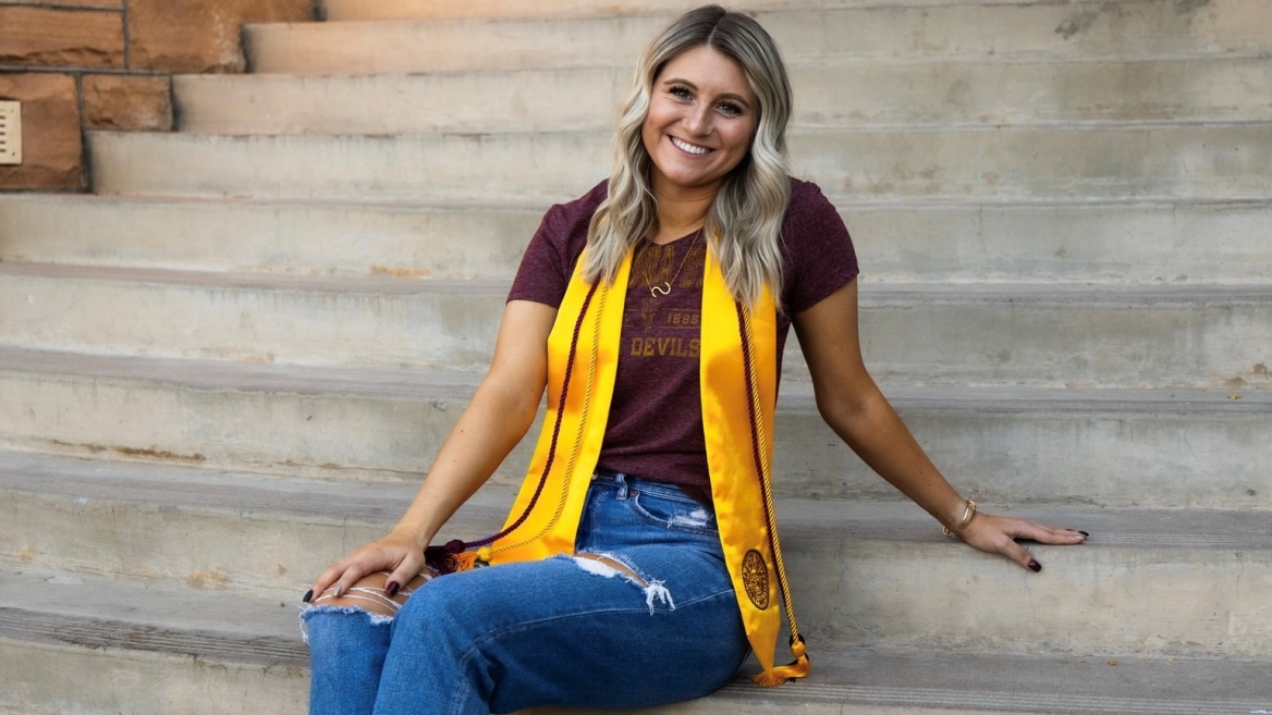 Sarah Bruder sits on concrete steps, smiling at the camera with her gold graduation sash and maroon cords draped around her neck